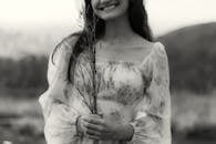 Black and white photo of a smiling woman holding wildflowers in a floral dress outdoors.